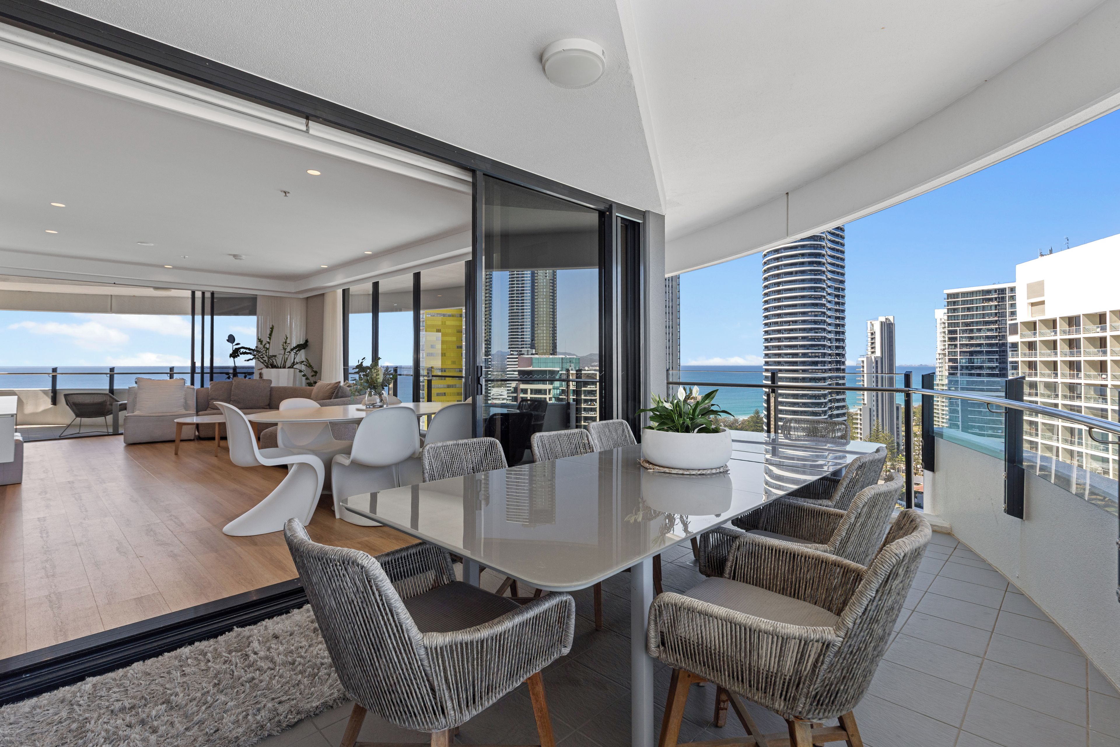 A modern living room with a glass dining table, wicker chairs, and a view of the ocean and city skyline.
