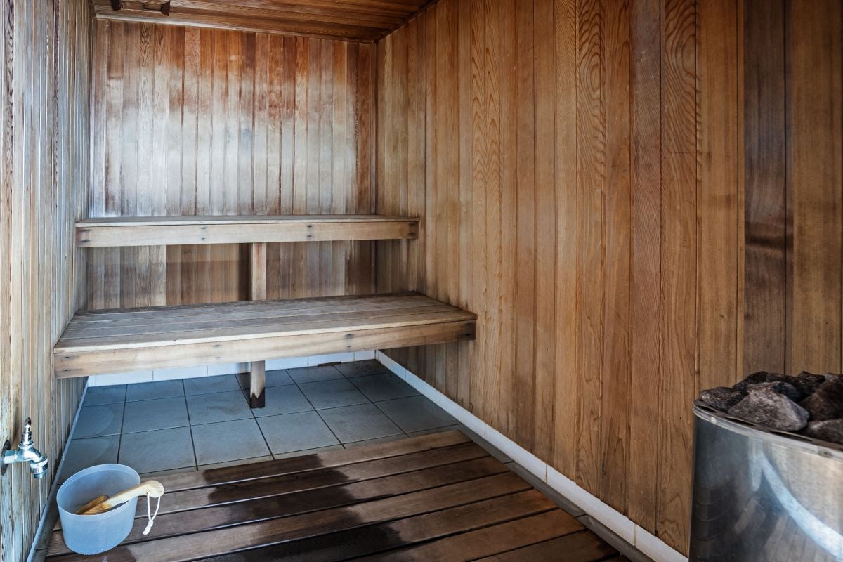 Una sauna con bancos de madera, un recipiente metálico con piedras y un cubo con asa de madera.