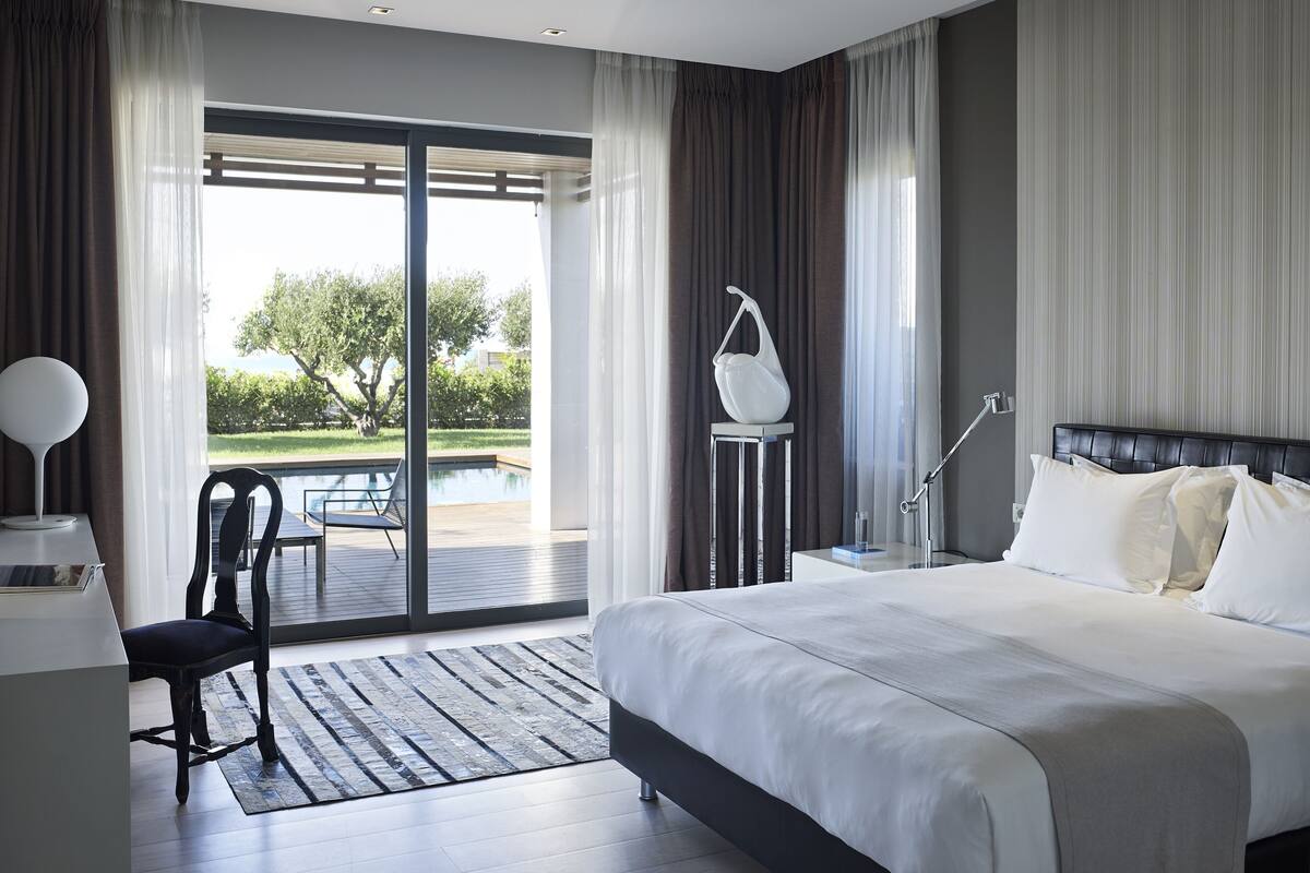 A modern hotel room with a large bed, a desk with a chair, a nightstand, and a view of a pool and greenery outside.