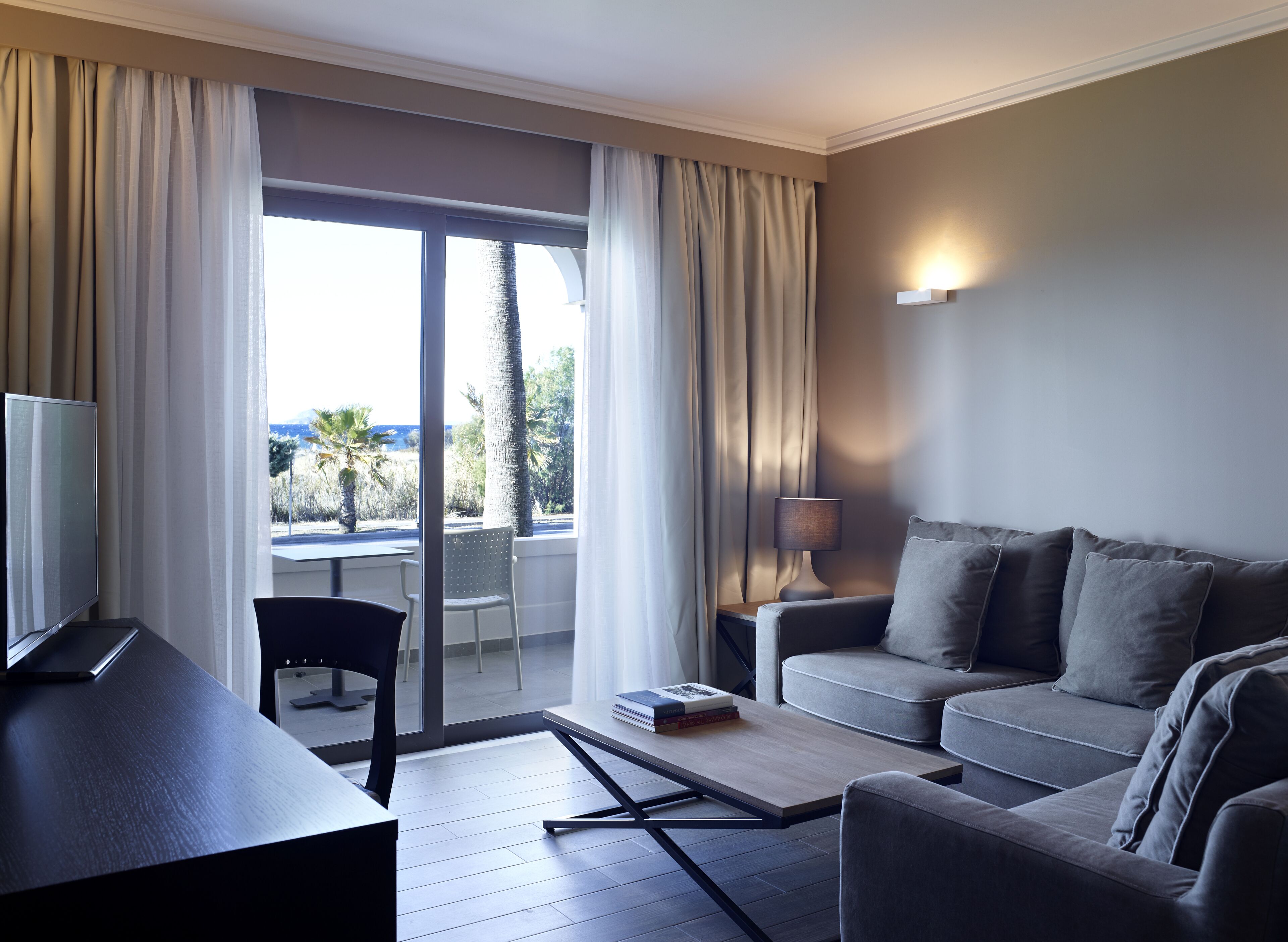 Habitación de hotel moderna con sofá, escritorio, lámpara y vistas a palmeras y montañas a través de amplias ventanas.