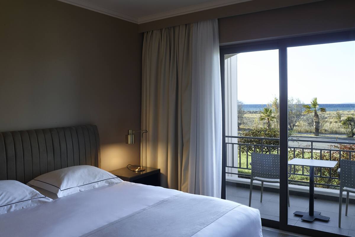 A hotel room with a bed, a nightstand, a lamp, and a view of the outdoors through a large window.