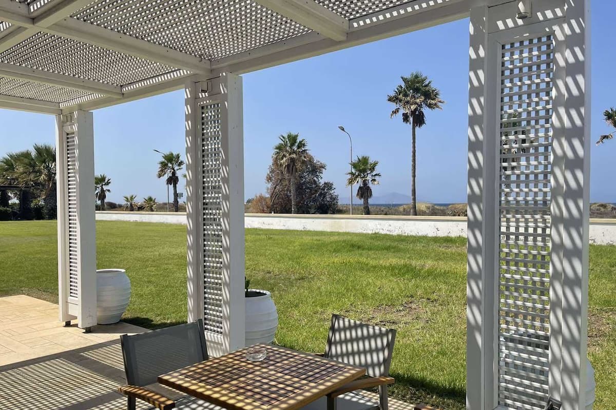 A covered outdoor seating area with a table and chairs, a white pergola structure, and a view of the beach and palm trees.
