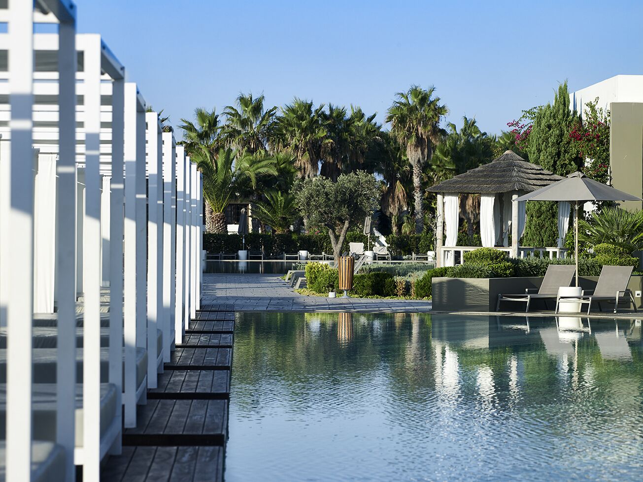Un área de piscina con muelle de madera, sillones de descanso, palmeras y un pabellón.