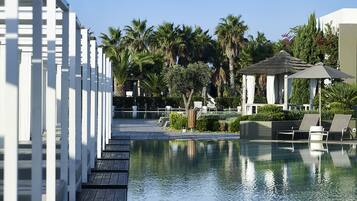 Un área de piscina con muelle de madera, sillones de descanso, palmeras y un pabellón.