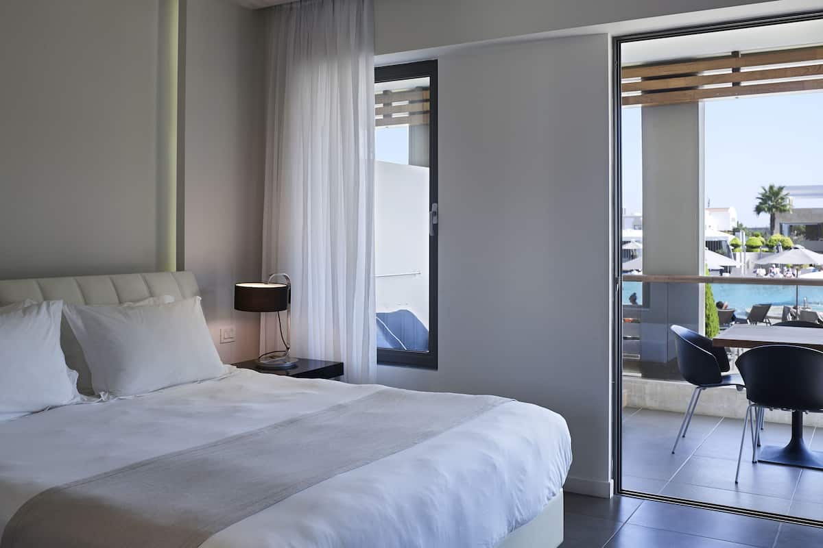 A hotel room with a bed, a nightstand, a lamp, a window with curtains, and a view of an outdoor pool area.