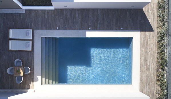 A modern outdoor pool with a minimalist design, surrounded by a wooden deck and a seating area with two lounge chairs and a table. - Pelagos Suites Hotel & Spa (Kos)