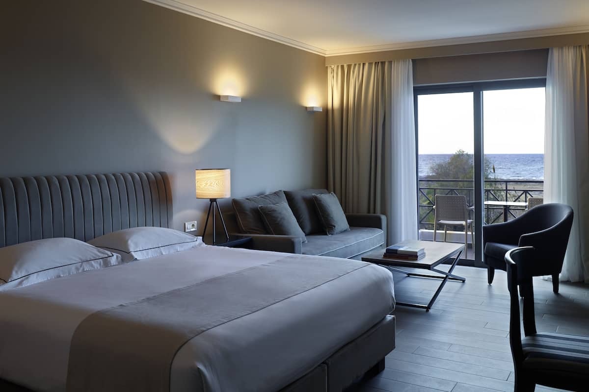 A hotel room with two beds, a sofa, a chair, a lamp, and a balcony with a view of the sea.