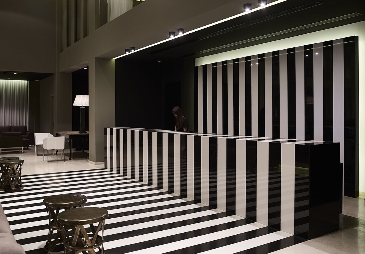 A modern lobby with a black and white striped floor, a seating area with tables and chairs, and a reception desk with a person standing behind it.