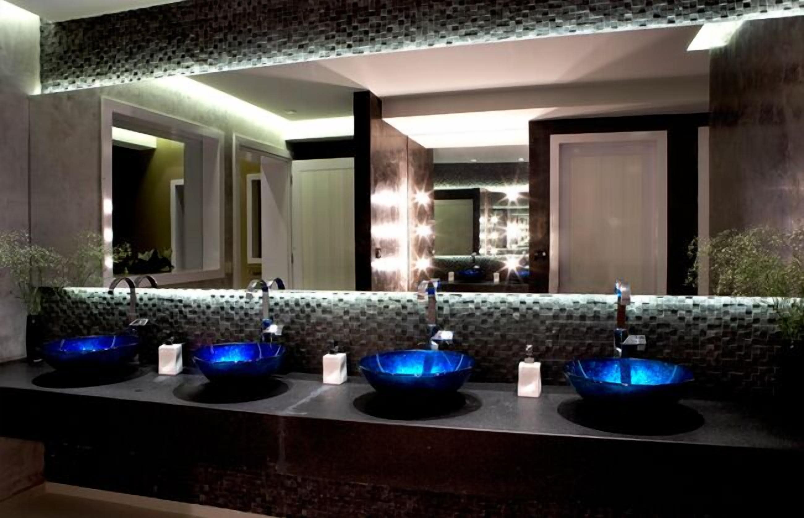 Un baño con tres lavamanos de cristal azul, un espejo y un respaldo de azulejos.