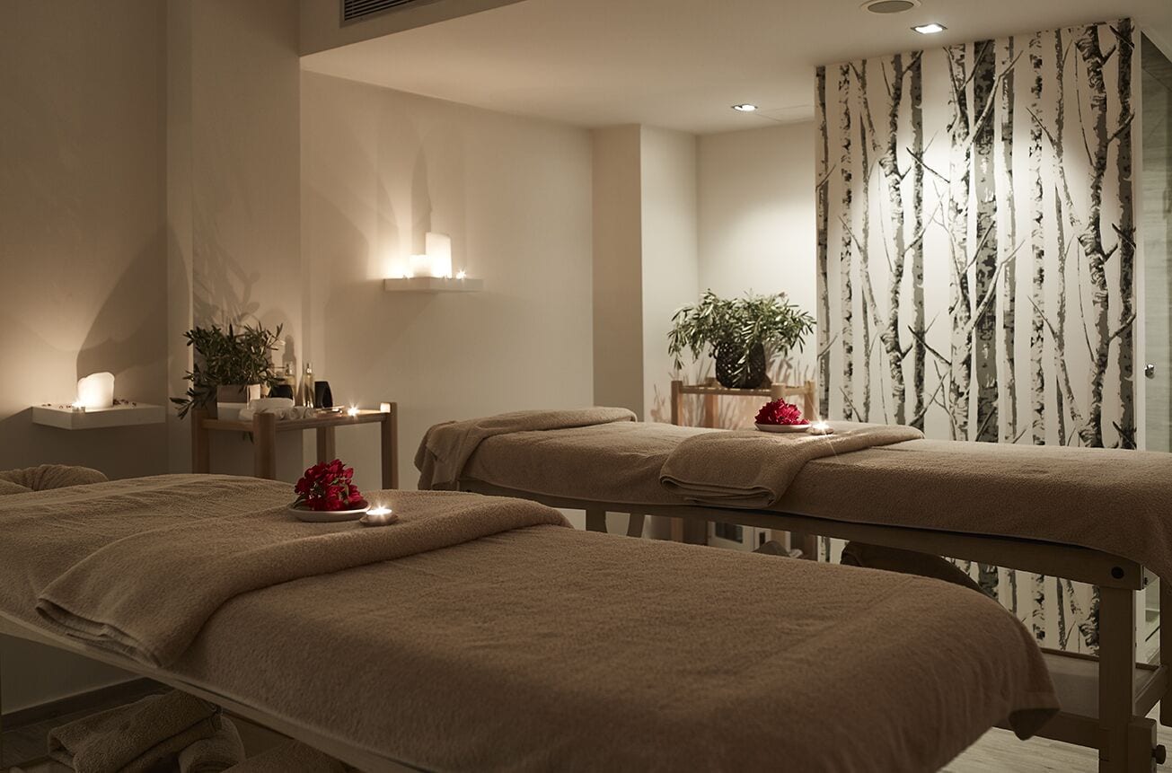 A spa room with two massage tables, a wall-mounted light fixture, and a decorative plant.