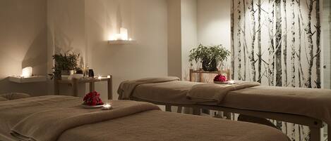 A spa room with two massage tables, a wall-mounted light fixture, and a decorative plant.