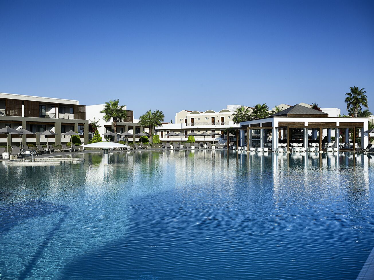 A large swimming pool with lounge chairs and umbrellas, surrounded by hotel buildings with balconies and palm trees.