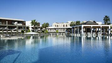 A large swimming pool with lounge chairs and umbrellas, surrounded by hotel buildings with balconies and palm trees.