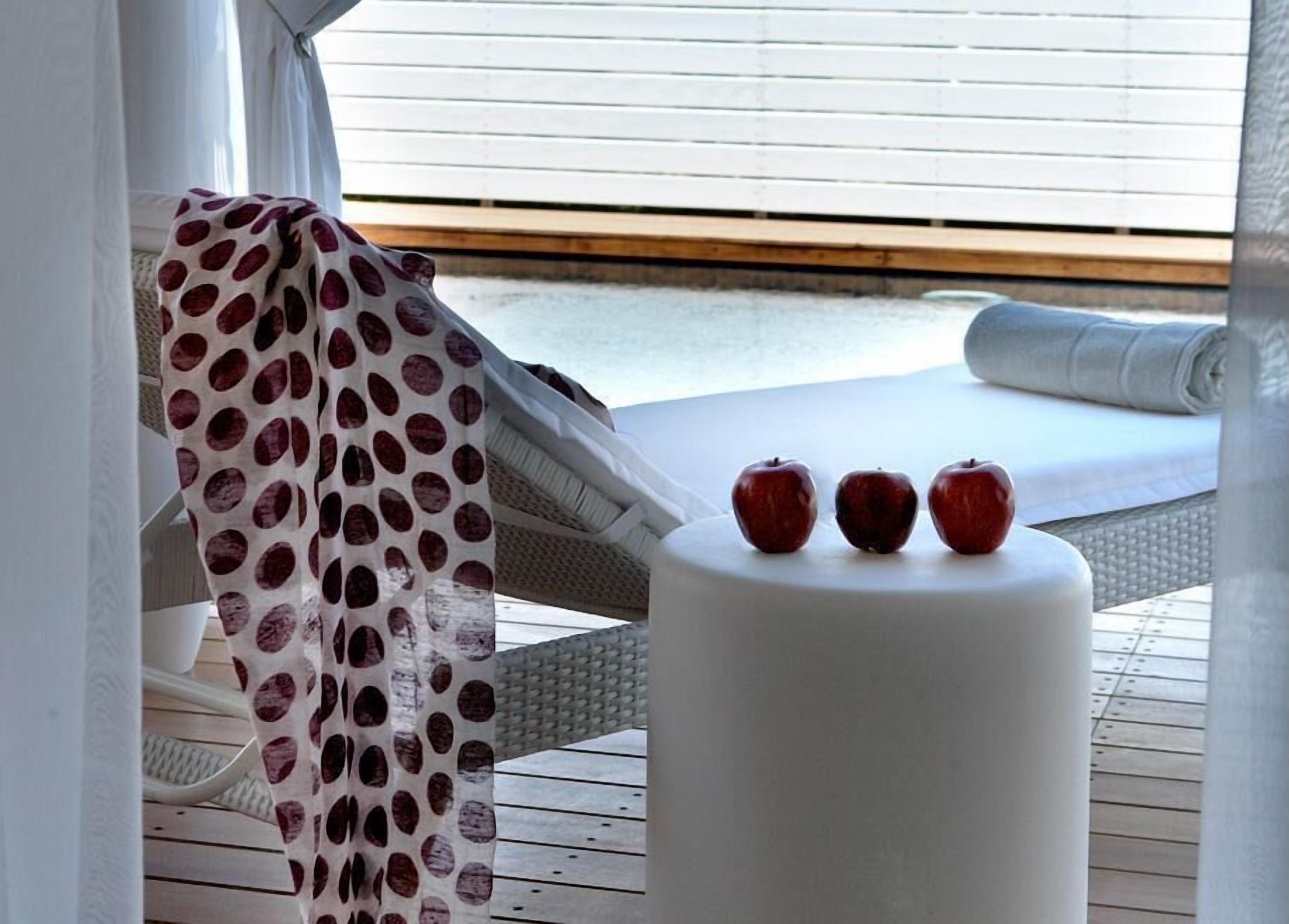 Una tumbona con una toalla de lunares, una mesa blanca con tres manzanas, y vista a la playa desde la ventana.