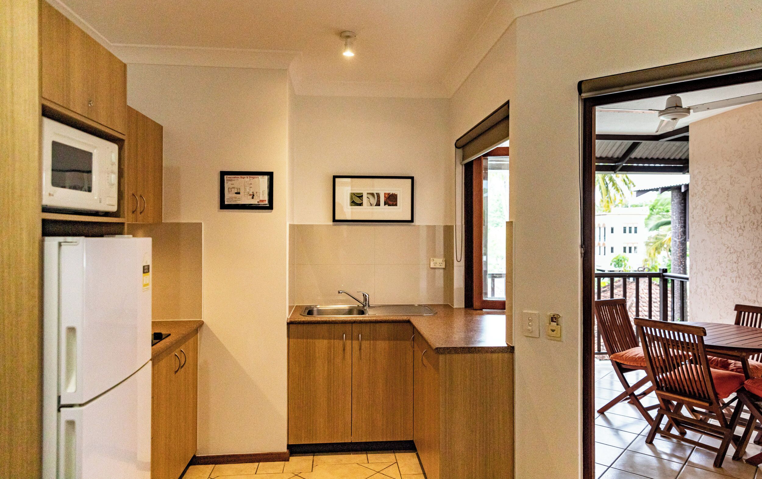 A compact kitchen with a refrigerator, microwave, and sink. There are framed pictures on the wall and a view of the outside through an open door.