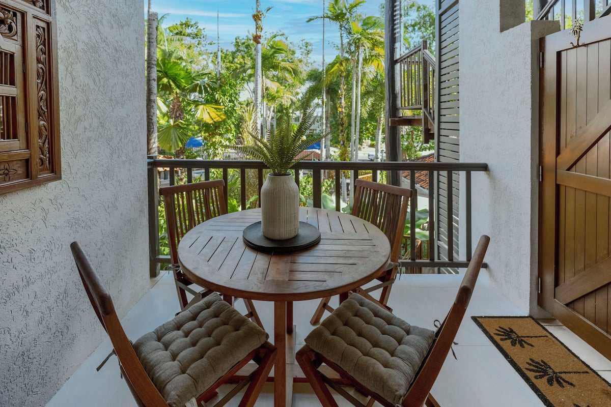 A round wooden table with two cushioned chairs, a potted plant, and a view of palm trees and a clear sky.
