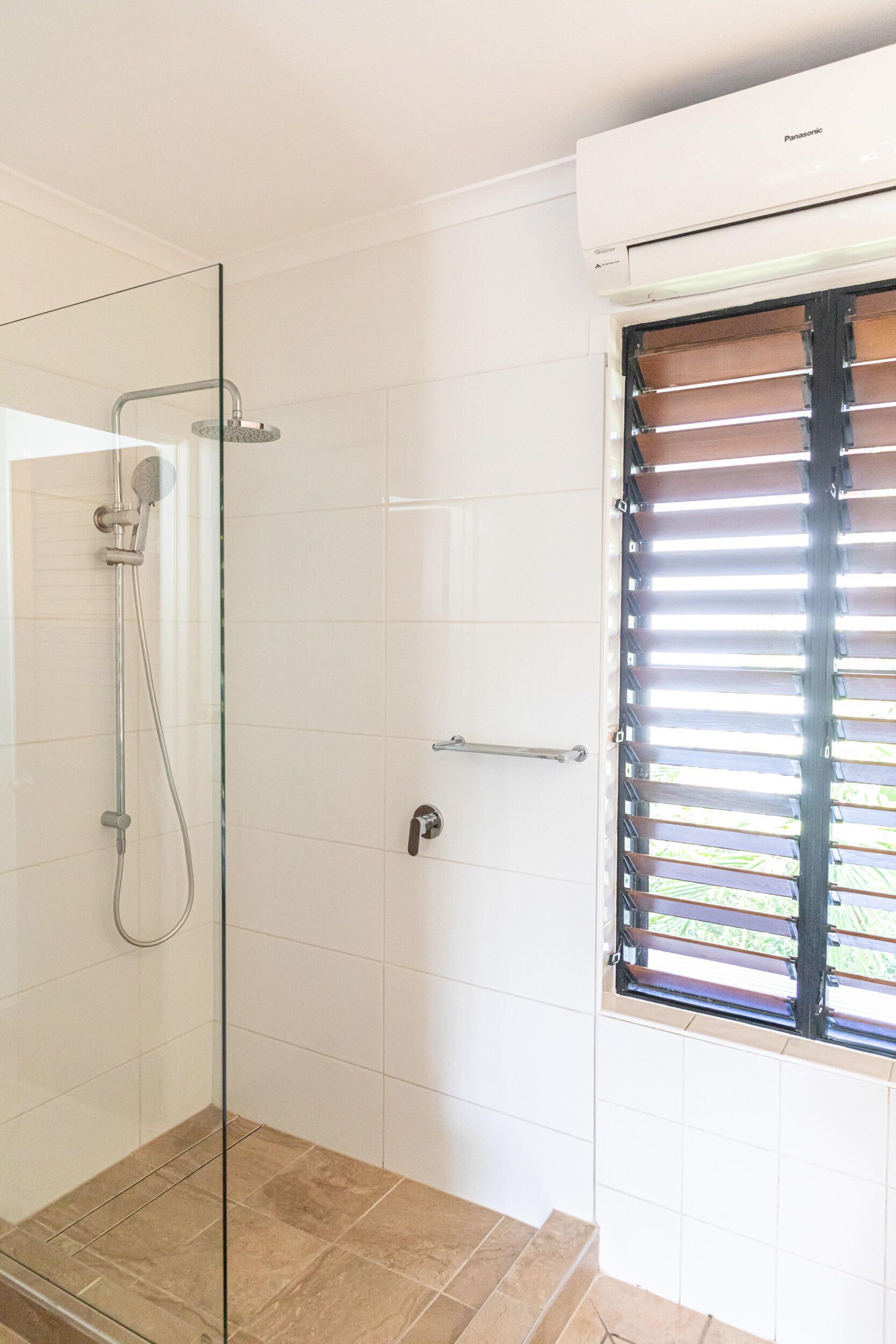 A bathroom with a glass shower enclosure, white tiled walls, a window with blinds, and a visible air conditioning unit.