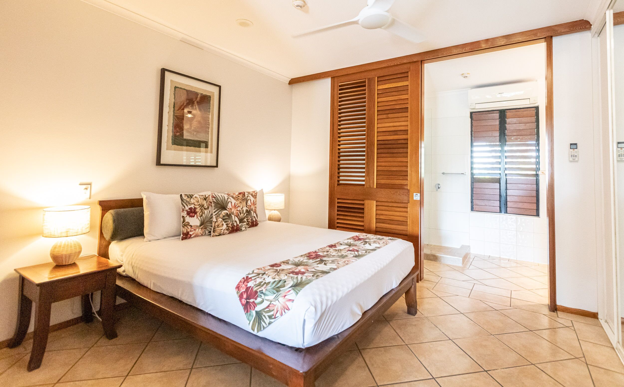 Un dormitorio con una cama, una mesita de noche, una lámpara, un cuadro enmarcado en la pared, una puerta de madera y un piso de baldosas.