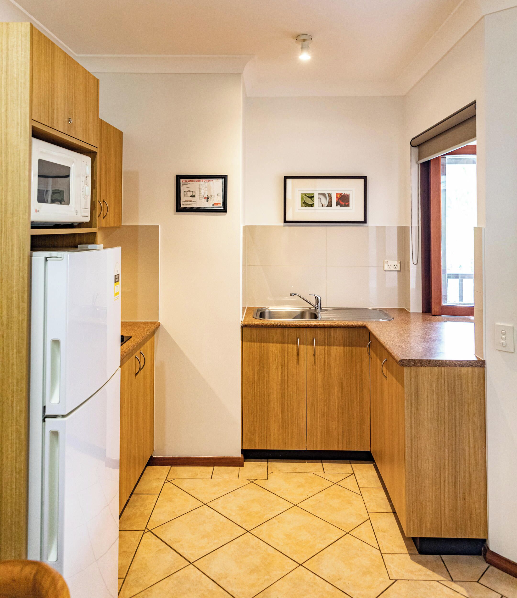 A compact kitchen with wooden cabinets, a built-in microwave, a sink, and a tiled floor.