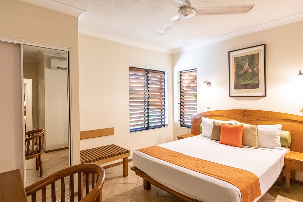 A hotel room with a large bed, wooden headboard, two windows with blinds, a bedside table, a chair, and a small bench.