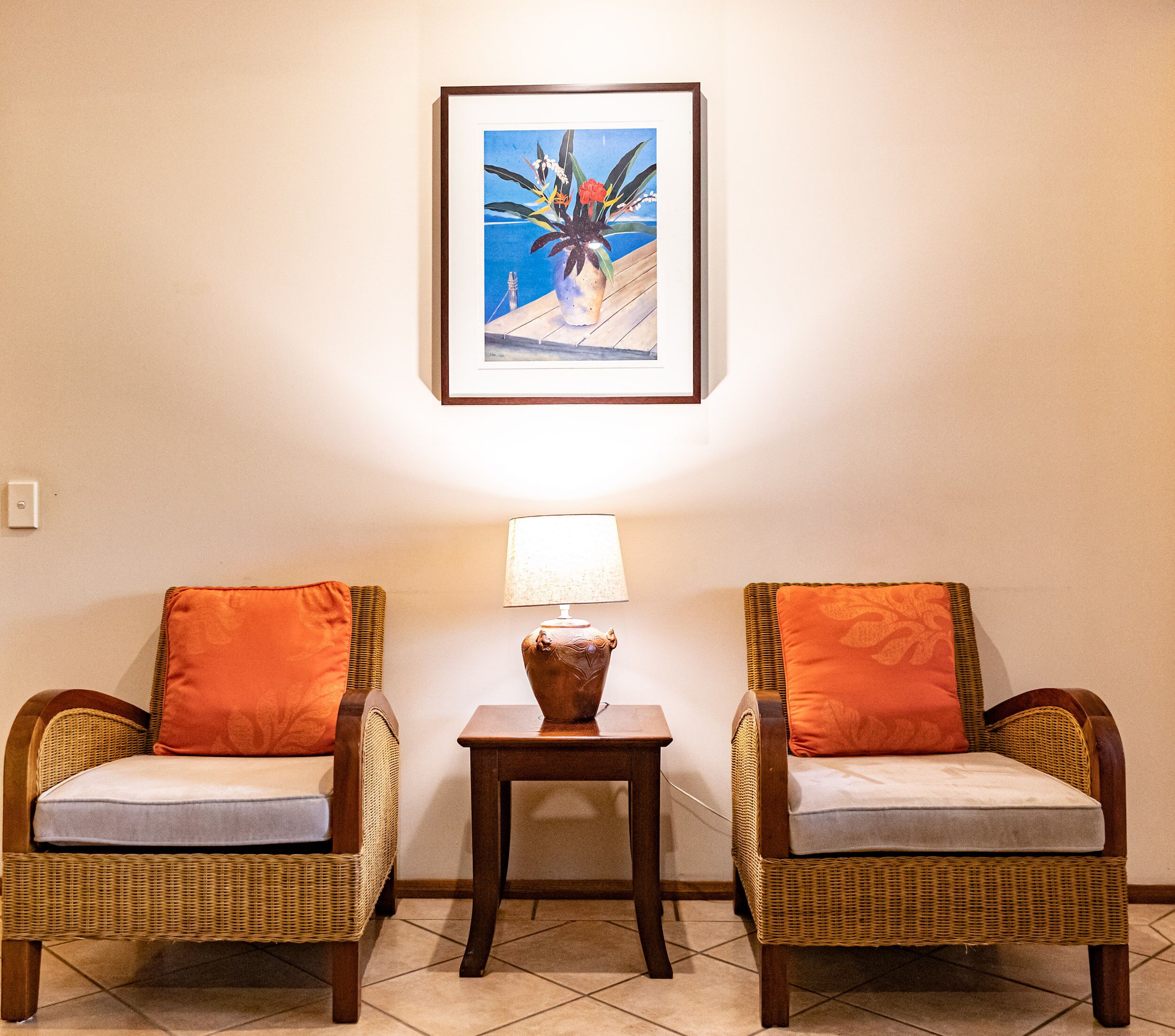 Deux fauteuils en osier avec des coussins orange, une petite table avec une lampe et un tableau encadré accroché au mur.