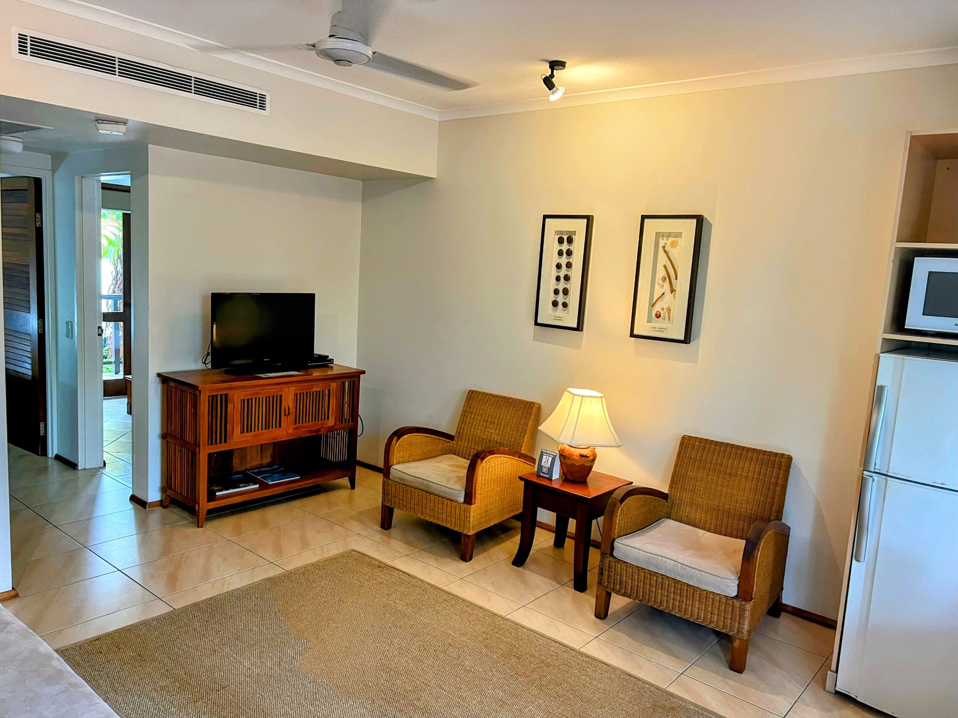 A living room with a television, two wicker chairs, a small table with a lamp, and a refrigerator.