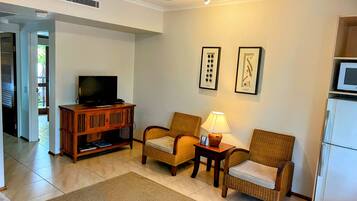 A living room with a television, two wicker chairs, a small table with a lamp, and a refrigerator.