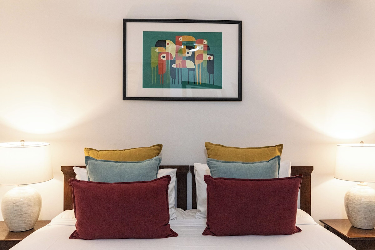 A neatly made bed with a variety of pillows, two bedside lamps, and a framed abstract painting on the wall.