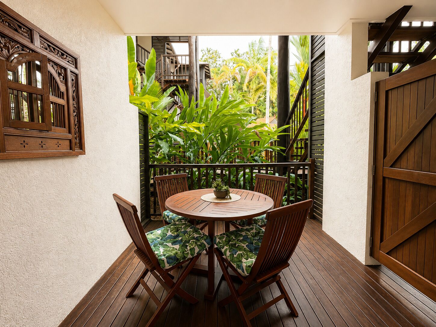 A wooden deck with a round table and chairs, surrounded by lush tropical plants.