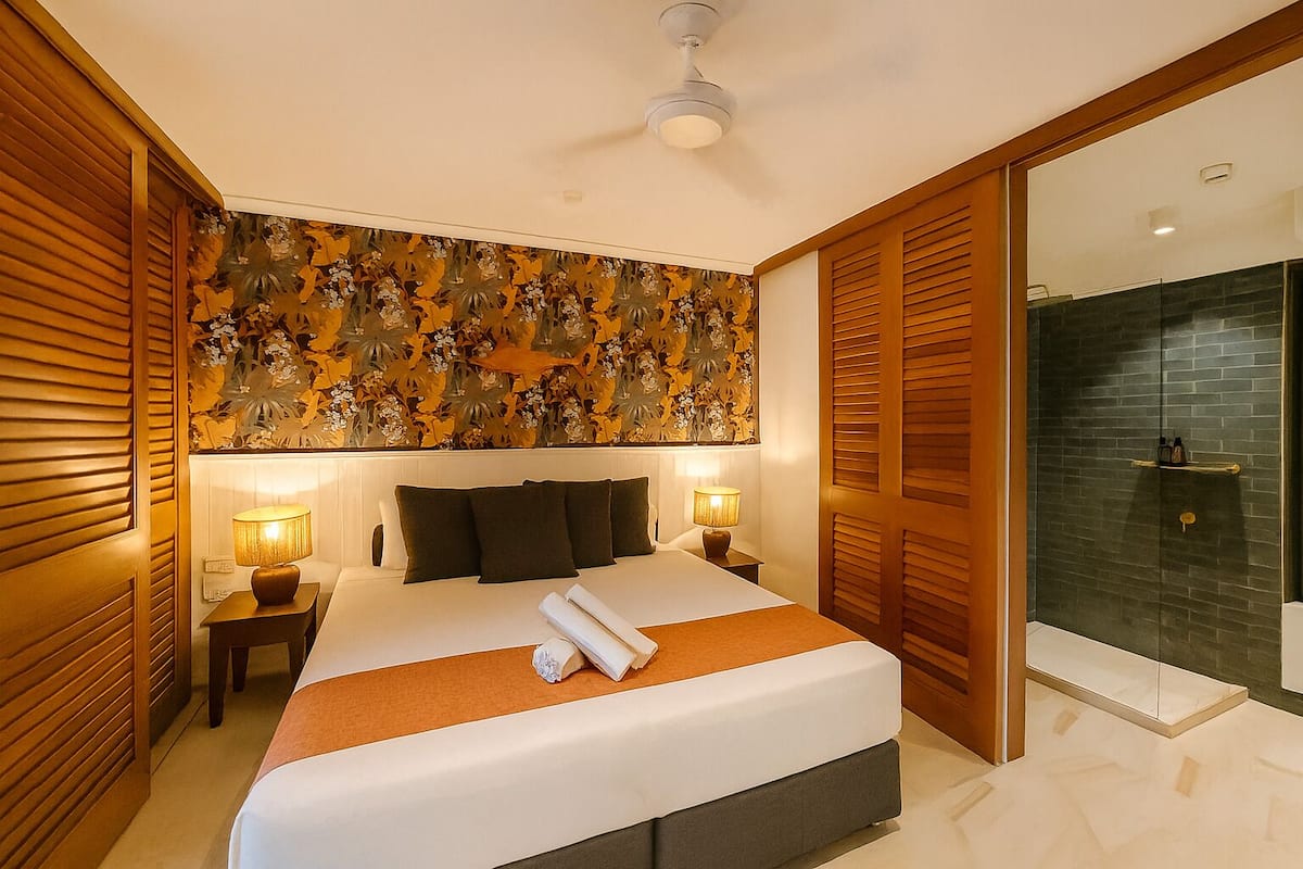 A hotel room with a large bed, two bedside lamps, a ceiling fan, and a shower area with a glass door.