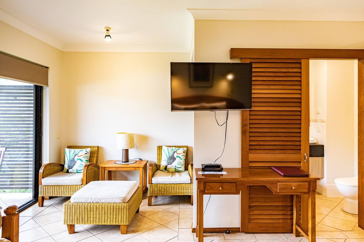 A modern hotel room with wicker furniture, a flat-screen TV, a desk, and a bathroom visible through an open door.