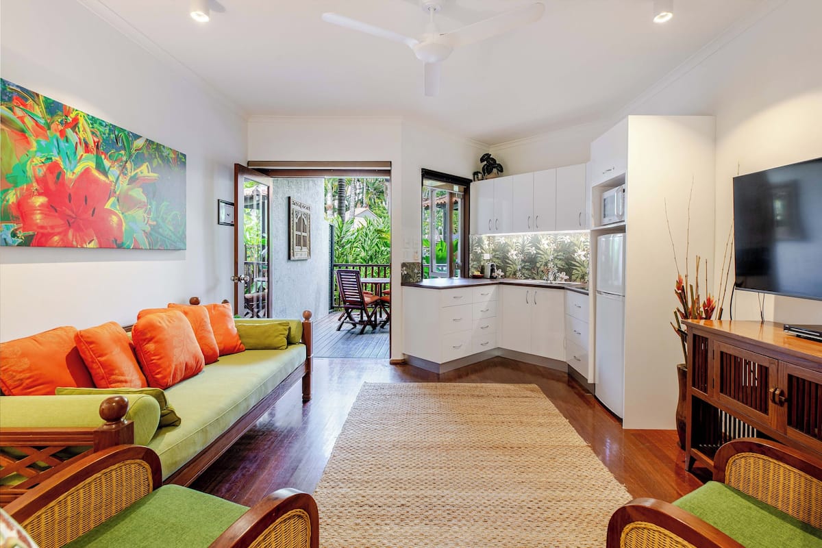 A modern living room with a green sofa, wooden chairs, a dining area, and a kitchen with white cabinets.