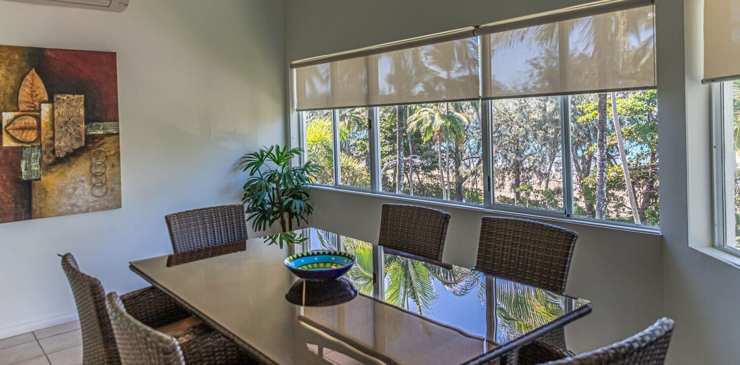 A dining area with a glass table, wicker chairs, and a view of tropical trees through large windows.