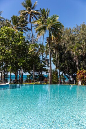 A clear blue pool surrounded by palm trees and other greenery, with a view of the ocean in the background. - Beaches Port Douglas (Port Douglas)