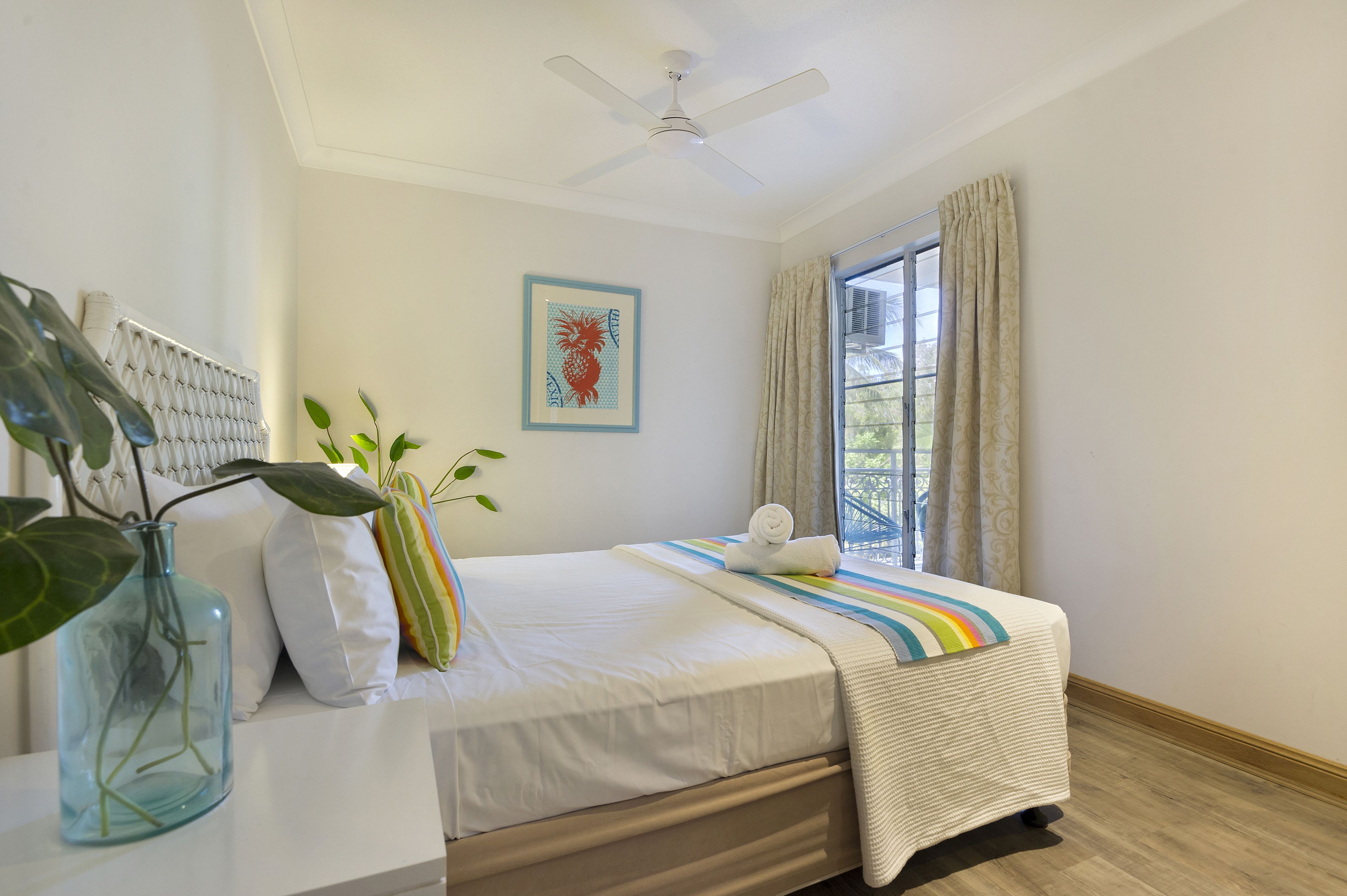 A neatly made bed with a striped blanket, a ceiling fan, a window with curtains, and a framed picture on the wall.