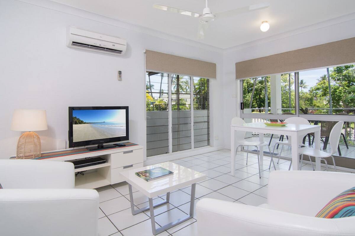 Una sala de estar moderna con un televisor de pantalla plana, muebles blancos y ventanas amplias que dan a zonas verdes.