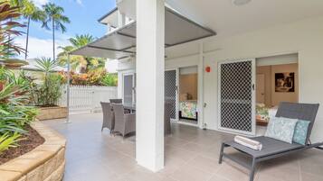A covered outdoor area with a dining table and chairs, a lounge chair with a towel, and a white fence with plants.