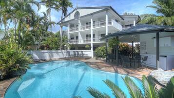 A two-story building with a swimming pool, surrounded by palm trees and a white fence.
