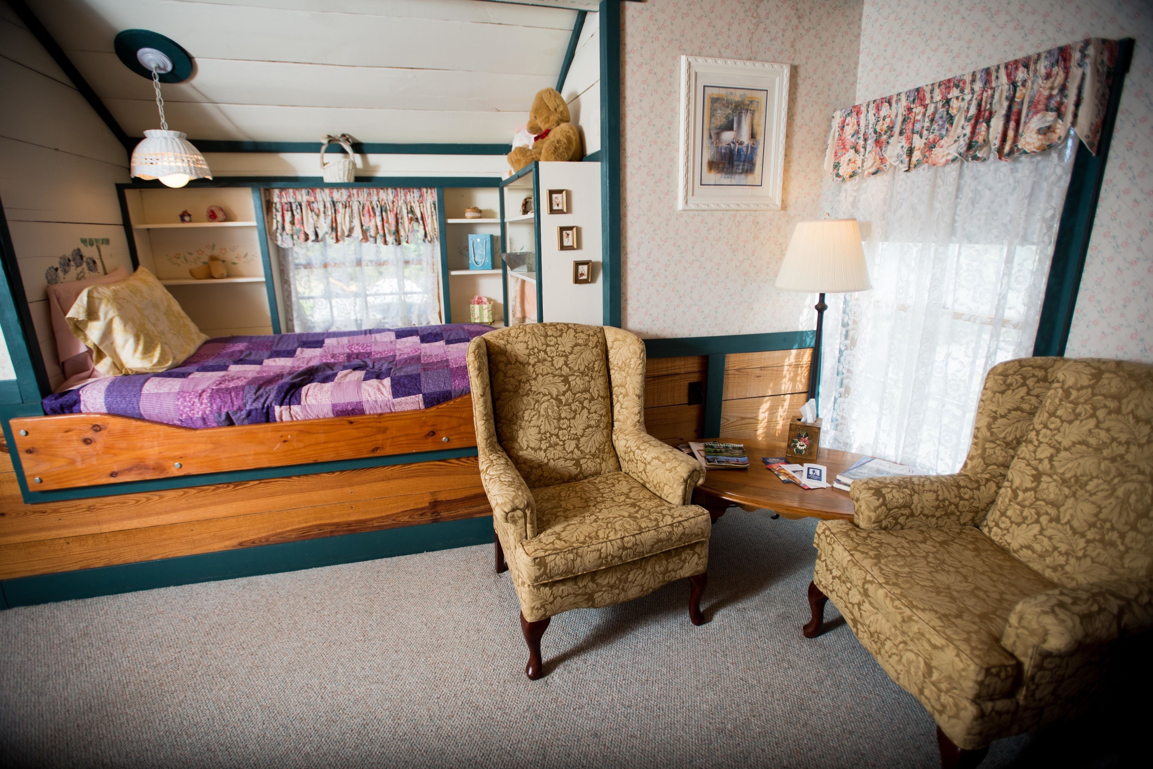 A bedroom with a wooden bed, two patterned armchairs, a small table, a lamp, a shelf with decorative items, and a window with curtains.