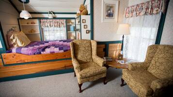 A bedroom with a wooden bed, two patterned armchairs, a small table, a lamp, a shelf with decorative items, and a window with curtains.