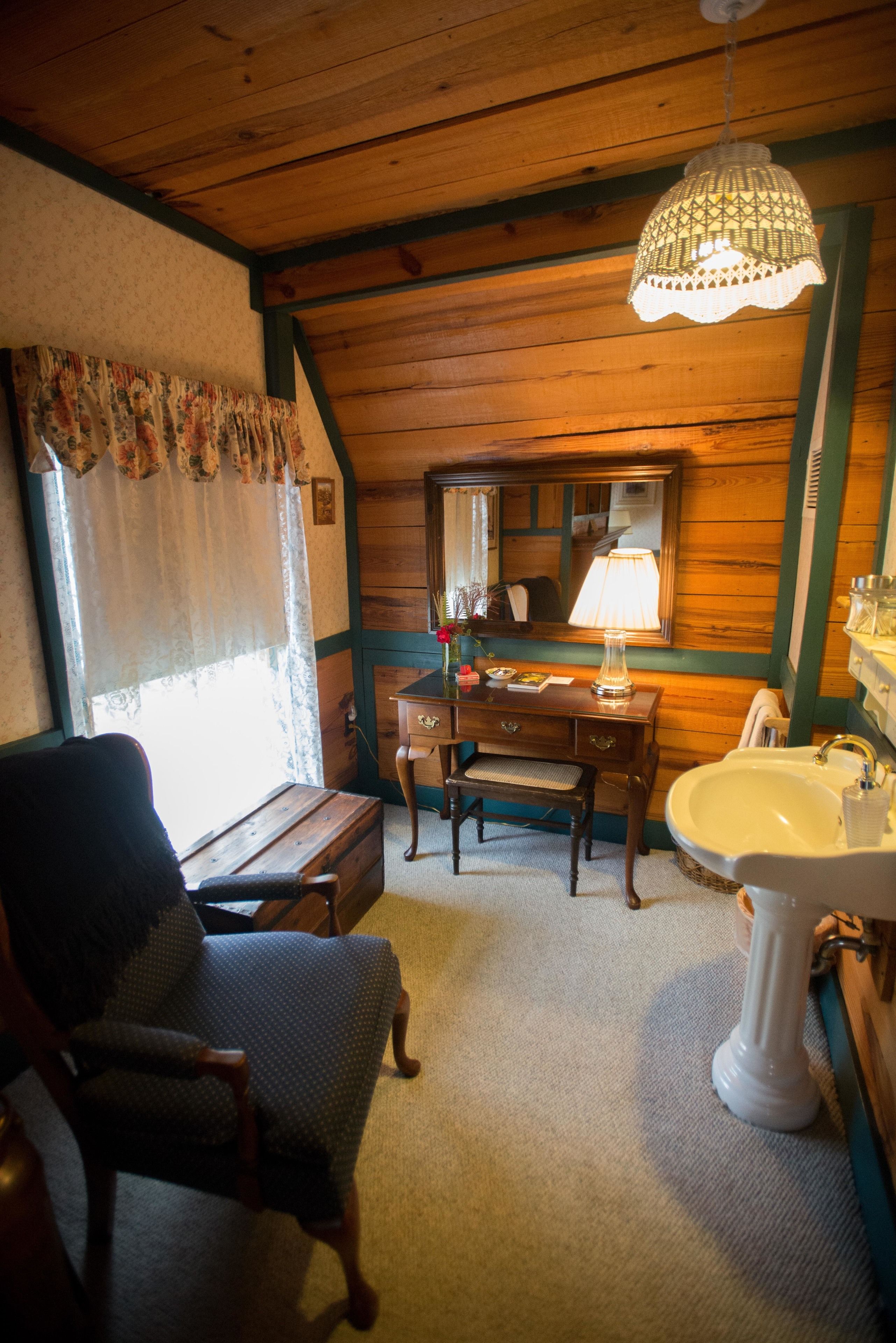A cozy room with a wooden ceiling, a bench, a chair, a dresser, a mirror, and a sink.