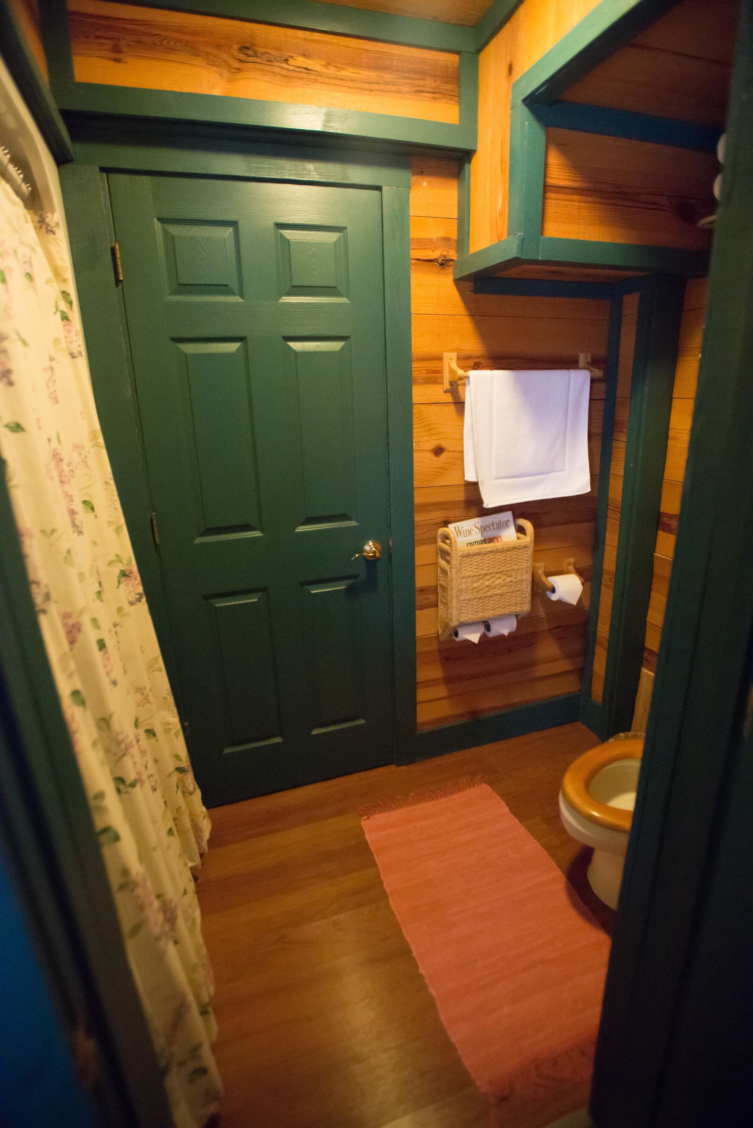 A small, wooden bathroom with a green door, a toilet, a wicker basket labeled "Wine Spectator," and a pink rug.