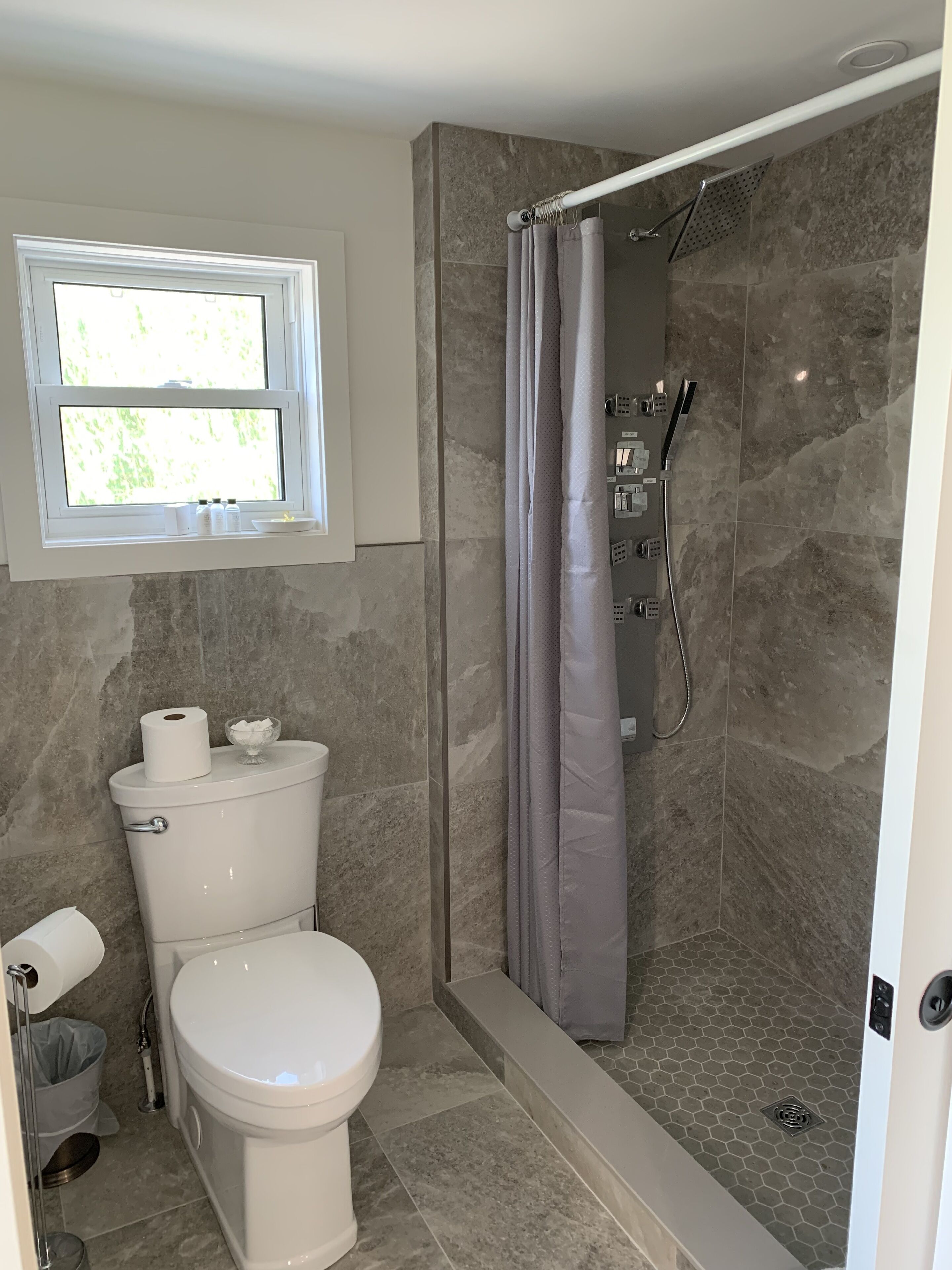A bathroom with a toilet, a shower with a grey curtain, and a window above the toilet.