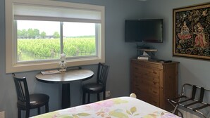 A room with a large window, a TV mounted on the wall, a wooden cabinet, and a table with chairs. - Green Oaks Bed and Breakfast (Niagara-on-the-Lake)