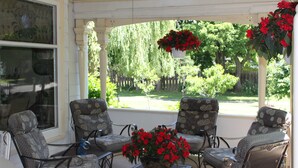 A covered patio with four patterned chairs, a table, and hanging flower baskets. - Green Oaks Bed and Breakfast (Niagara-on-the-Lake)