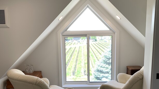 A room with a sloped ceiling, a large window with a view of a green landscape, and two beige armchairs. - Green Oaks Bed and Breakfast (Niagara-on-the-Lake)