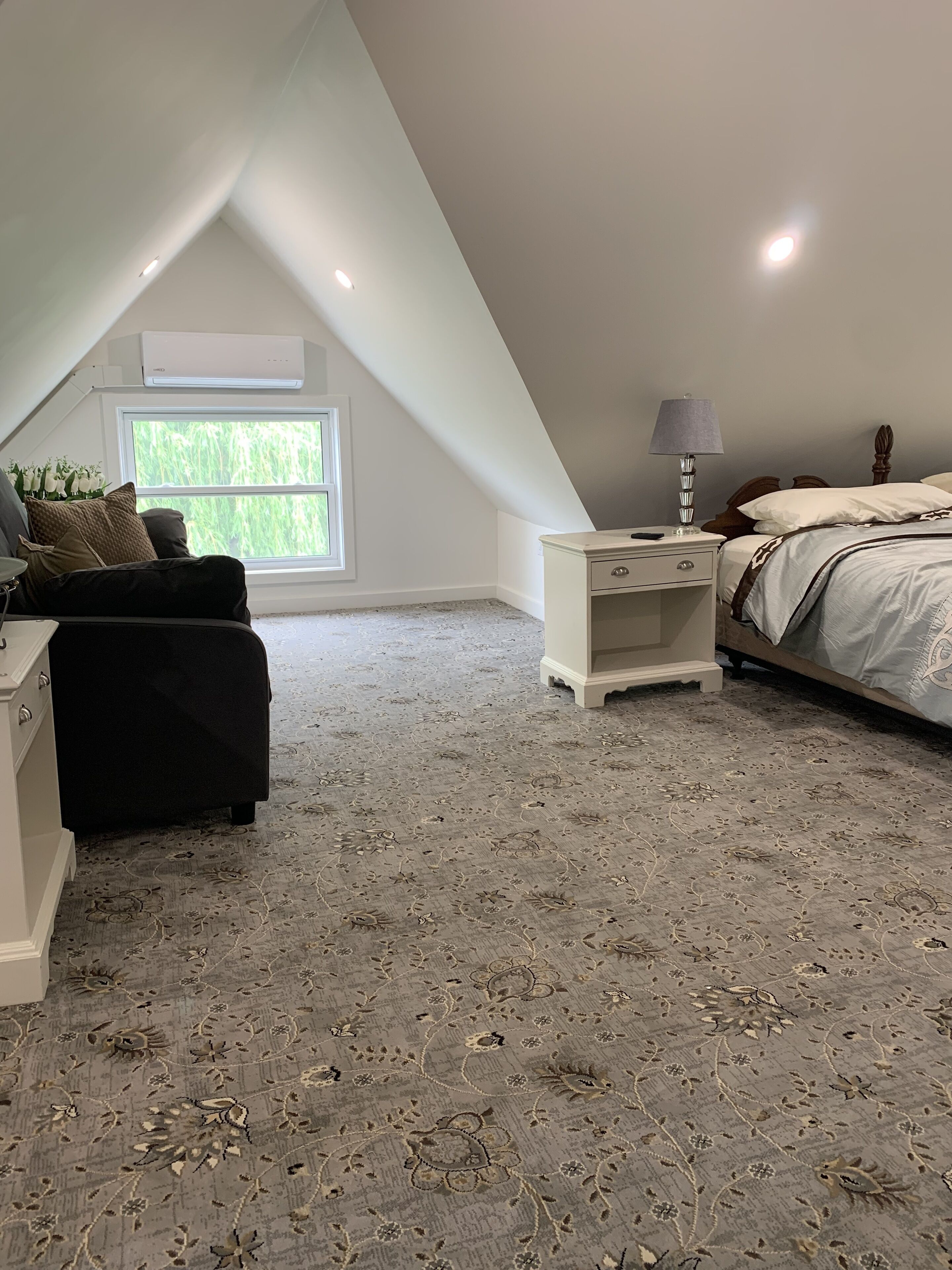 A bedroom with a sloped ceiling, a bed with a patterned bedspread, a nightstand with a lamp, a dresser, and a window with a view of greenery.