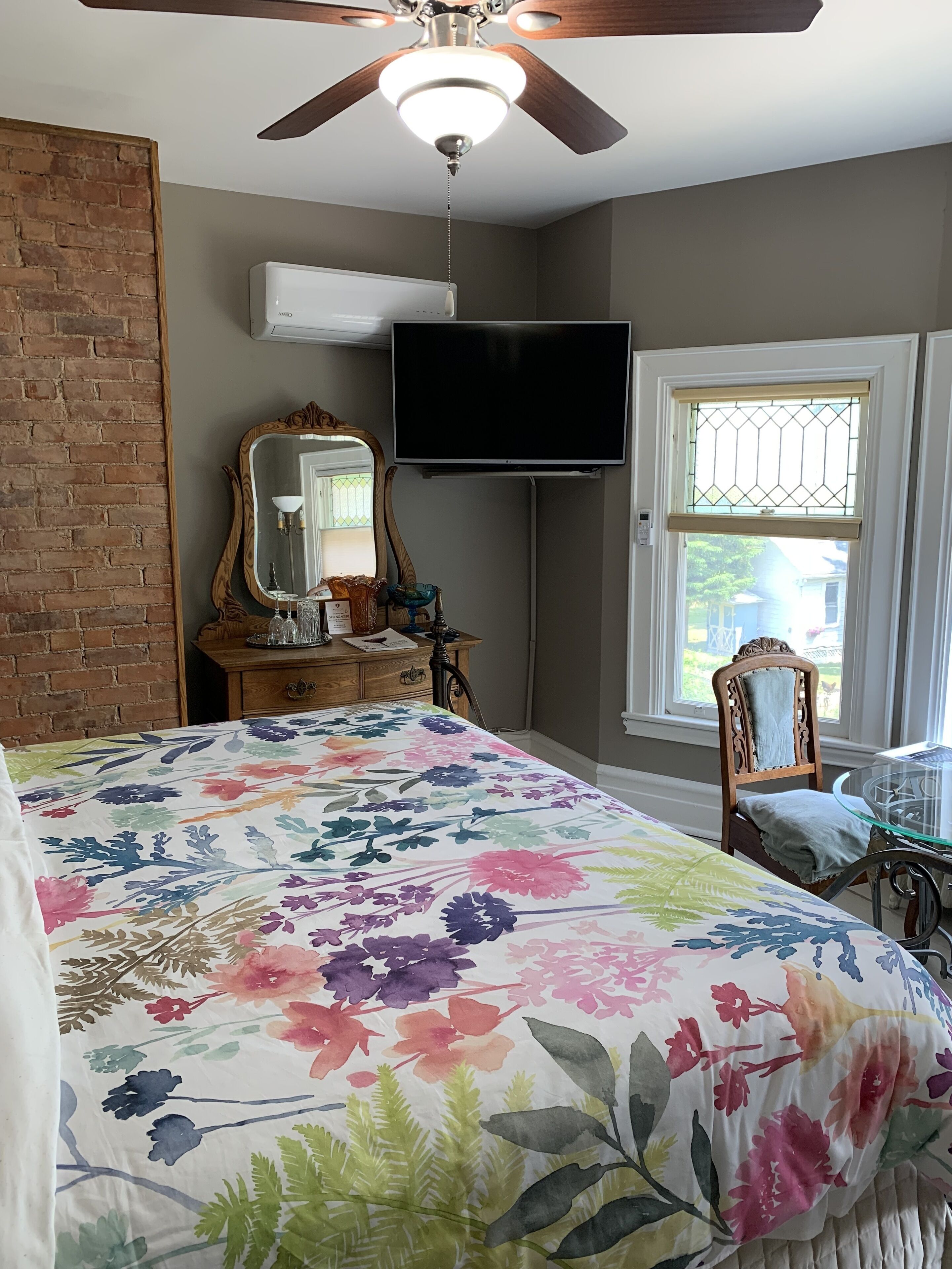 A bedroom with a large floral bedspread, a ceiling fan, a flat-screen TV, a dresser, a mirror, and a window with a view of the outside.