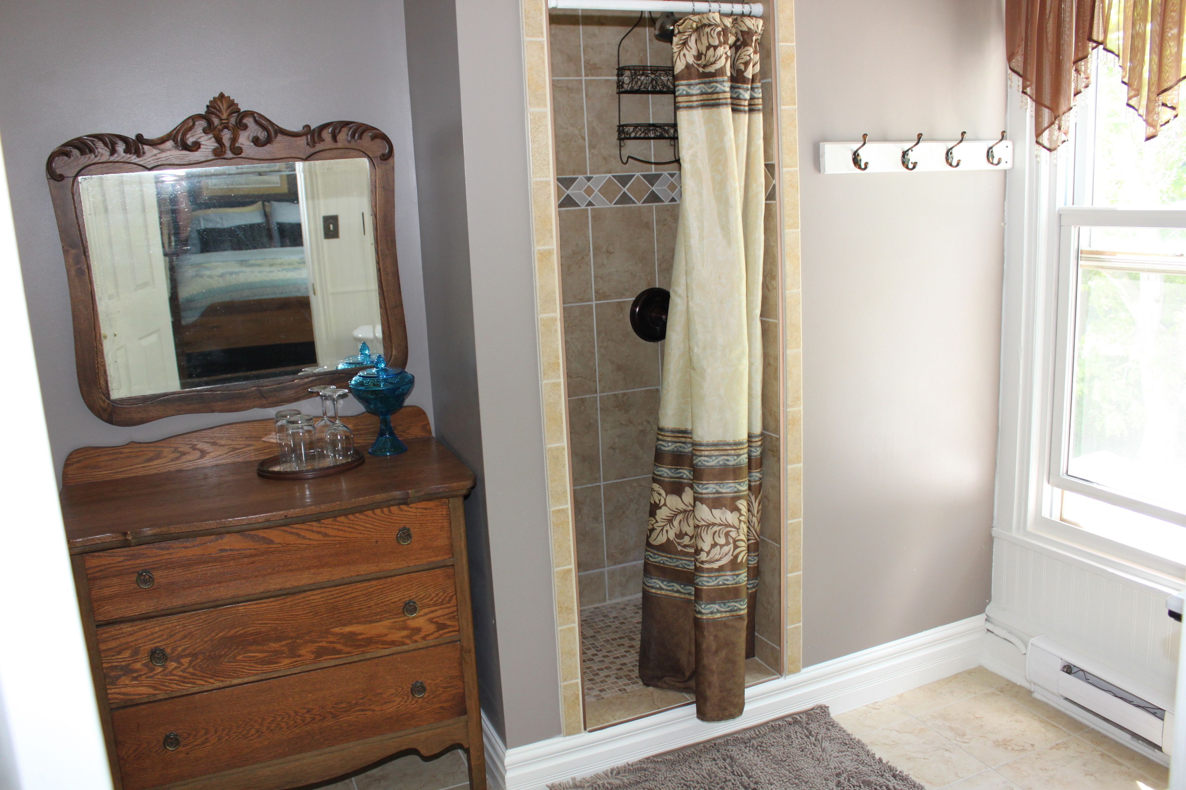 Une salle de bain avec une commode en bois, une douche avec un rideau à motifs, un miroir et une fenêtre également dotée d’un rideau.