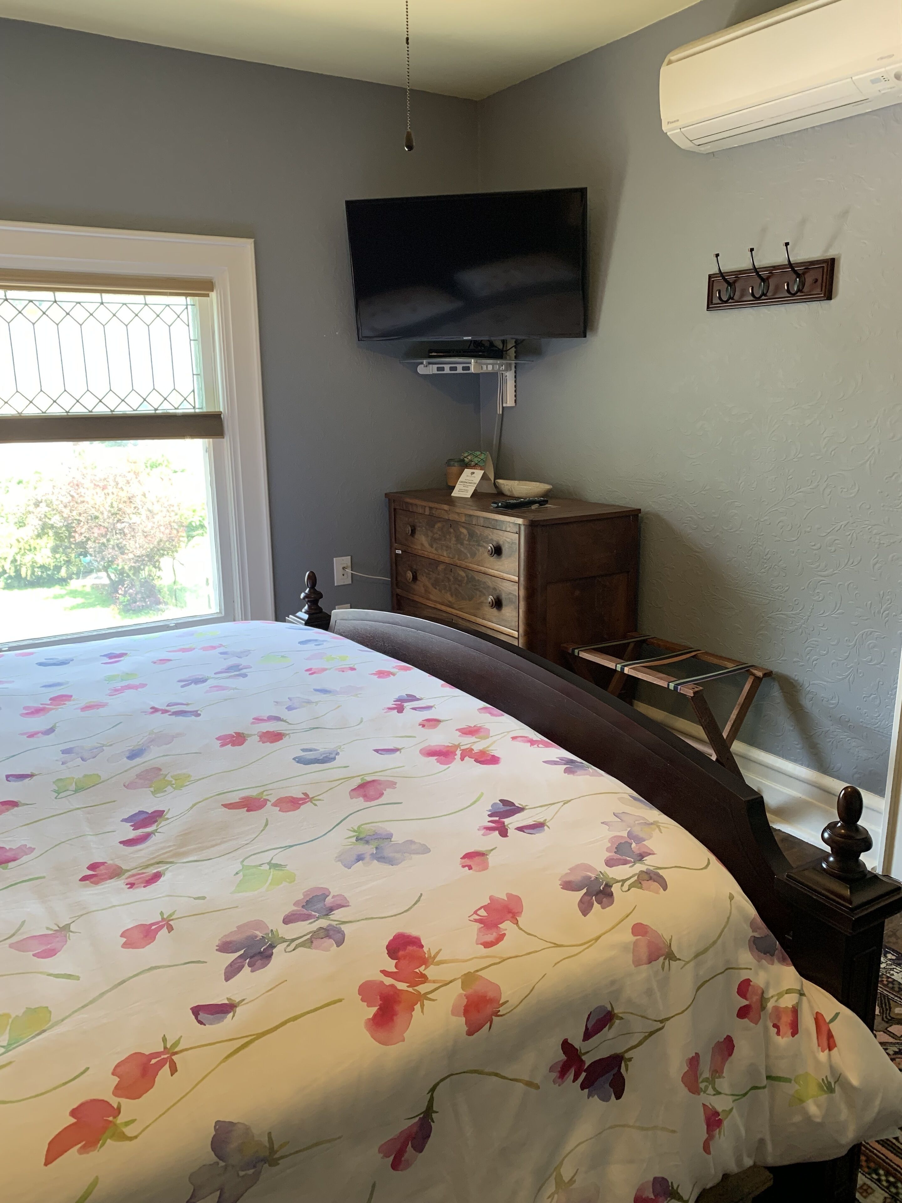 A bedroom with a floral bedspread, a television mounted on the wall, a wooden dresser, and a window with a view of greenery outside.